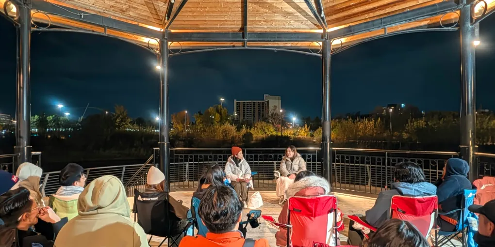 People gather at night in Calgary’s open-sided Music Pavilion while two people sit at the center to share stories